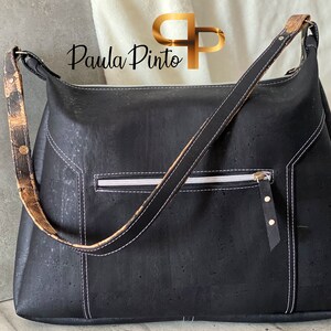 May include: A black cork handbag with a long brown and black strap. The bag has a white zippered pocket and gold accents.