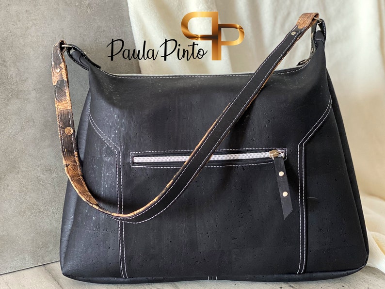 May include: A black cork handbag with a long brown and black strap. The bag has a white zippered pocket and gold accents.