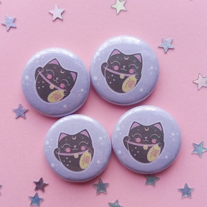 Può includere: Quattro bottoni rotondi, lilla chiaro, con un'illustrazione di un gatto nero. Il gatto ha una luna crescente sulla testa, un collare rosa e tiene in mano una moneta d'oro con testo giapponese. Lo sfondo è lilla chiaro con puntini e stelle bianche.