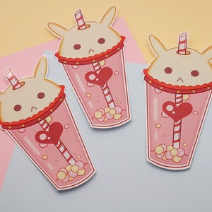 Puede incluir: Tres pegatinas que representan una bebida rosa con una cara de conejo. La bebida tiene un corazón rojo y una pajita a rayas. La bebida está decorada con pequeñas burbujas redondas amarillas y rosas.