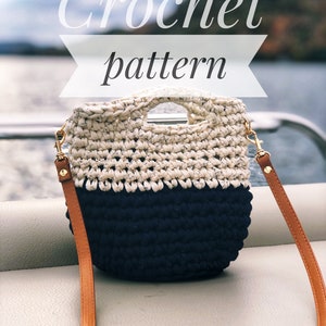 May include: Crochet pattern for a white and navy blue tote bag with a brown strap. The bag has a top handle and a detachable strap.
