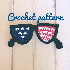 May include: Crochet pattern for a pair of sunglasses made with red, white and blue yarn. The sunglasses are displayed against a white wooden background.