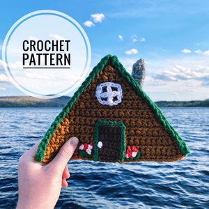 May include: A crocheted cabin with a brown exterior, green trim, and a white door. The cabin has a white crocheted window with a cross design. The cabin is being held in front of a blue lake and a blue sky.