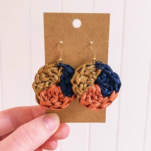 May include: A pair of woven earrings with a circular design. The earrings are made of three colors: brown, orange, and blue.