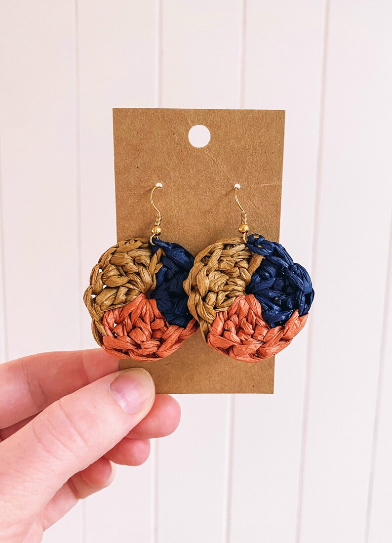 May include: A pair of woven earrings with a circular design. The earrings are made of three colors: brown, orange, and blue.