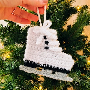 May include: White crocheted ice skate ornament with black trim and black buttons. The ornament is hanging on a green Christmas tree.