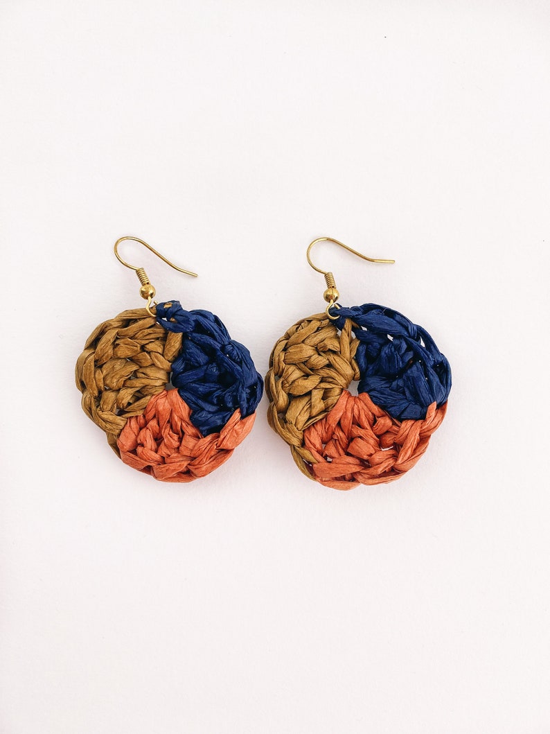 May include: A pair of handmade earrings with a circular design. The earrings are made of woven straw in shades of brown, orange, and blue.