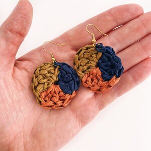 May include: A pair of woven circle earrings with gold hooks. The earrings are made of three colors: brown, orange, and dark blue.