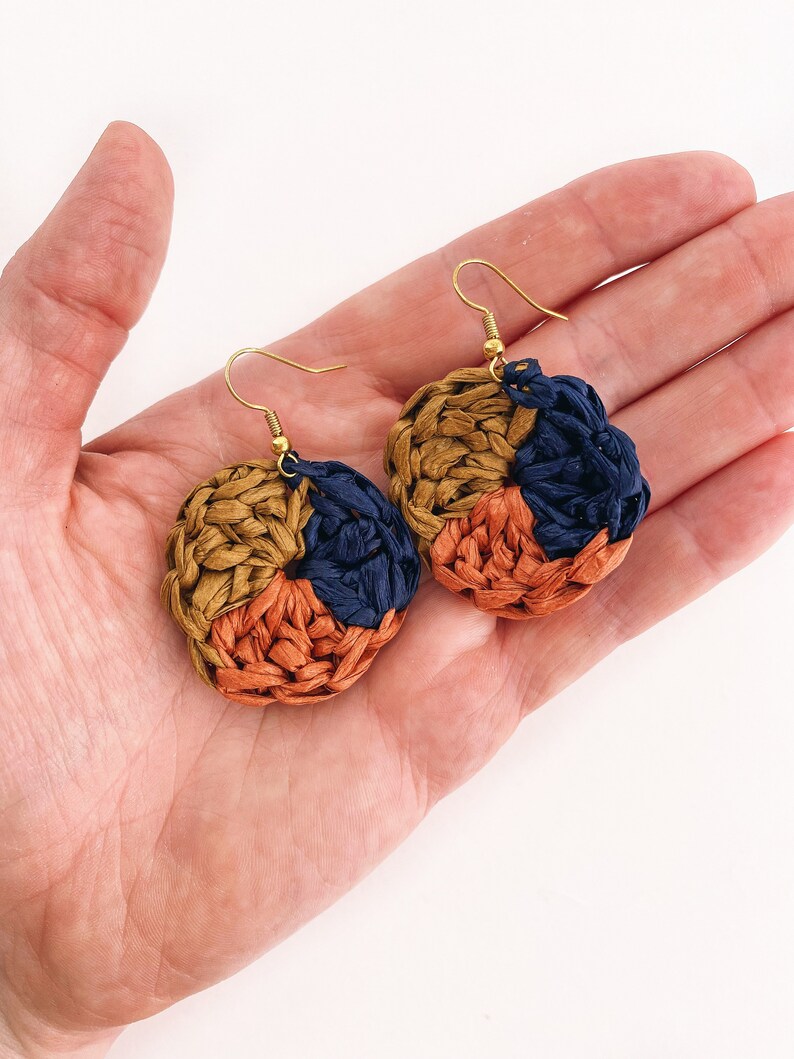 May include: A pair of woven circle earrings with gold hooks. The earrings are made of three colors: brown, orange, and dark blue.