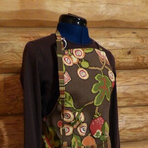 May include: A brown apron with a floral print featuring brown, green, red, and white flowers and berries. The apron has a large pocket with a striped fabric lining.