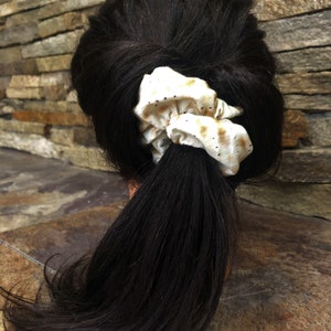 May include: A close-up of a ponytail with a cream-colored scrunchie. The scrunchie has a pattern of small brown spots. The hair is dark brown and the scrunchie is tied around the base of the ponytail.