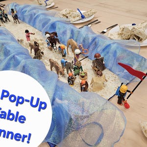 May include: A table runner with a blue, shimmering fabric design, featuring a 3D pop-up scene with miniature figures and animals. The text "3D Pop-Up Table Runner!" is in a white circle. The table is set with plates and silverware.