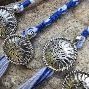 May include: Three silver charm bracelets with blue and white braided cords. Each charm features a Hebrew word in a decorative font.