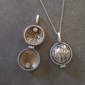 Puede incluir: Un collar de plata con un colgante de medallón que presenta una escena de montaña detallada con una luna creciente y estrellas. El medallón se abre para revelar un interior en blanco.