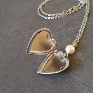 Puede incluir: Un collar de plata con un dije en forma de corazón y un dije de perla. El dije está abierto, revelando un interior vacío.