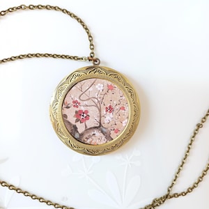 以下が含まれることがあります： 花柄の金色のロケットネックレス。ロケットは繊細なチェーンに付いており、ピンクとブラウンの色の花柄が描かれた丸いペンダントが付いています。