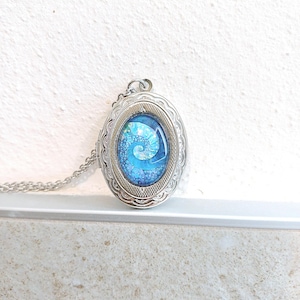 Op de afbeelding: Zilverkleurige ovale hangerketting met een blauw-wit spiraalontwerp. De hanger heeft een decoratieve rand en hangt aan een delicate ketting. De ketting wordt gepresenteerd tegen een witte achtergrond.