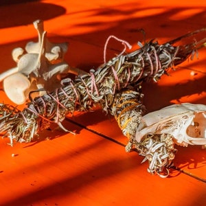 Könnte beinhalten: Nahaufnahme von drei Gegenständen auf einer roten Oberfläche: ein weißer Tierschädel, ein Bündel getrockneter Salbei, das mit rosa Schnur gebunden ist, und ein weiteres Bündel getrockneter Salbei, das mit gelber Schnur gebunden ist.