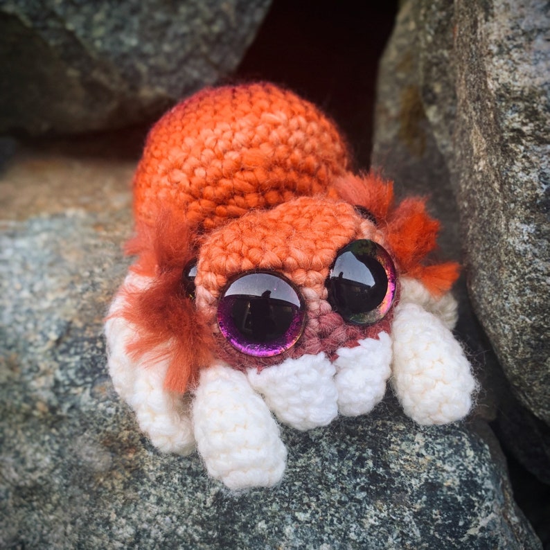 May include: A crocheted plush toy shaped like a spider with a red and orange body and white legs. The spider has large, round, purple eyes.