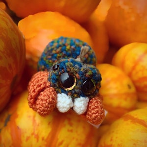 May include: A crocheted spider toy with large, colorful eyes. The spider is sitting on a pile of orange and yellow tomatoes.