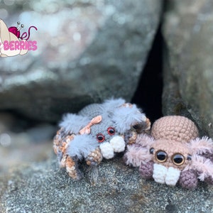 May include: Two crocheted spider plushies, one gray with a pink bow and the other brown, are sitting on a rock surface. Both plushies have large, black eyes and white teeth.