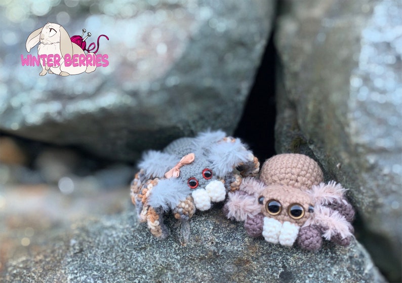 May include: Two crocheted spider plushies, one gray with a pink bow and the other brown, are sitting on a rock surface. Both plushies have large, black eyes and white teeth.