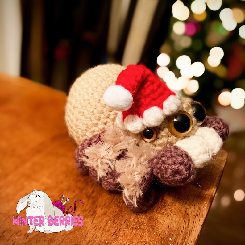 May include: A crocheted spider wearing a red and white Santa hat. The spider has large brown eyes and is sitting on a wooden surface.