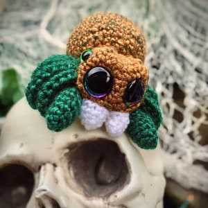May include: A crocheted spider toy with a brown body, green legs, and large iridescent eyes. The spider is sitting on a white skull.
