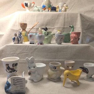 May include: A selection of vintage ceramic egg cups in assorted designs and colours. The egg cups feature animal and character designs, including a rooster, bunny, duck, and a dog. Some have painted details and are displayed on a white cloth.