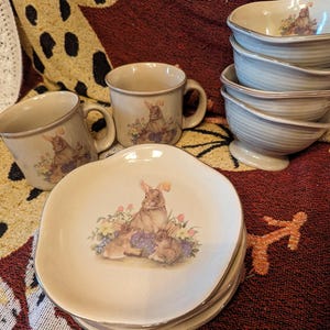 May include: Set of four beige ceramic mugs with a brown bunny rabbit design. The mugs are stacked on top of each other. There are also four beige ceramic bowls with a brown bunny rabbit design. The bowls are stacked on top of each other.