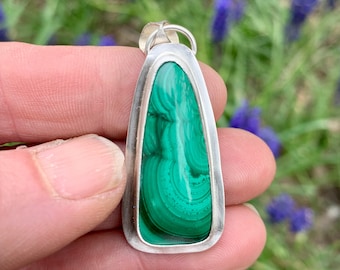 May include: A green malachite stone pendant set in a silver frame. The stone has a swirling pattern of green and black.