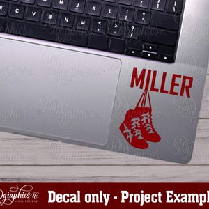 Op de afbeelding: Een rode vinyl sticker met de naam "MILLER" en twee rode schoenen die aan de letters hangen. De sticker zit op een zilveren laptop.