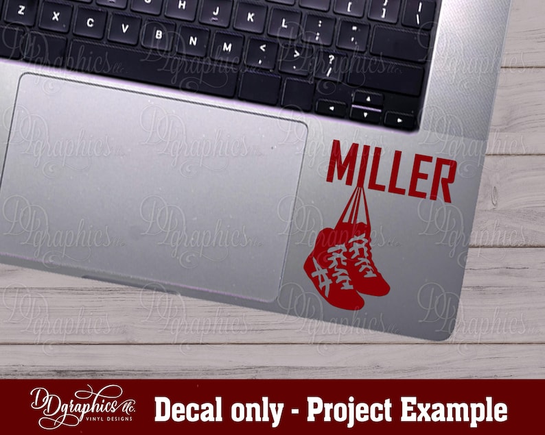 Op de afbeelding: Een rode vinyl sticker met de naam "MILLER" en twee rode schoenen die aan de letters hangen. De sticker zit op een zilveren laptop.
