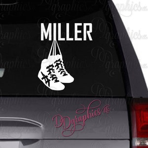 Op de afbeelding: Witte vinyl sticker met het woord "MILLER" en twee witte sneakers die aan de letters hangen. De sneakers zijn aan elkaar gebonden met een touwtje.