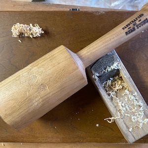 May include: A wooden mallet with a handle that reads "Handcrafted by -  Ryan Young". The mallet is sitting on a wooden surface with wood shavings.