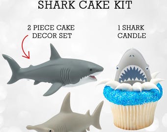 May include: Shark Cake Kit with a gray shark and a hammerhead shark figurine. Includes a cupcake with a shark candle and blue sprinkles. The text "SHARK CAKE KIT" is at the top of the image.