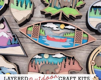 May include: A collection of layered wooden craft kits with outdoor themes. Designs include a canoe on a lake, a bear in a forest, and a deer in a forest. The text "LAYERED Outdoor CRAFT KITS" is displayed.