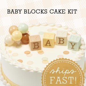 Op de afbeelding: Een wit geglazuurde taart versierd met baby-thema-elementen. De taart heeft vier vierkante blokken met de tekst "BABY" en een cluster van ronde, pastelkleurige decoraties. De tekst "BABY BLOCKS CAKE KIT" staat bovenaan.