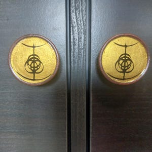 May include: Two circular cabinet knobs featuring a metallic gold finish on the front face. Each knob is decorated with a black, intricate, symmetrical line-art symbol centred on the gold surface. The knobs have a dark brown, wood-toned edge.