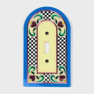 Puede incluir: Una cubierta decorativa para interruptor de luz con borde azul, centro amarillo y fondo a cuadros blanco y negro. La cubierta presenta un diseño decorativo con hojas verdes y corazones burdeos.