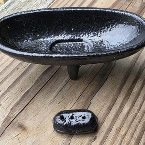 May include: An oval-shaped black soap dish with a textured finish, supported by three short legs, accompanied by a small, separate black rectangular piece featuring three circular holes aligned in a row on its top surface.