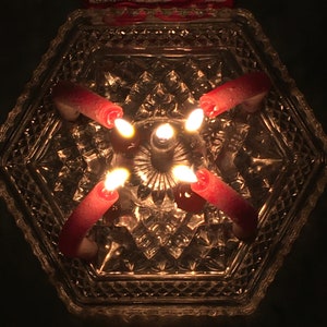 Pode incluir: Quatro velas vermelhas estão acesas dentro de um prato hexagonal de vidro transparente com um padrão de diamante. As velas estão dispostas em forma de cruz.