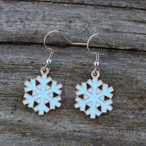 May include: A pair of snowflake earrings. Each earring features a light blue snowflake design with a gold-colored outline and silver-colored ear hooks. The earrings are displayed on a weathered wooden surface.