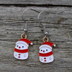 May include: A pair of Christmas-themed earrings. Each earring features a small, white snowman with a red hat and scarf. The snowman has black dot eyes and a gold-colored metal hook.