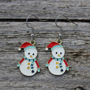 May include: A pair of snowman earrings. Each earring features a white snowman with a red hat, blue scarf, and red arms. The earrings have silver-colored hooks and are set against a weathered wooden background.