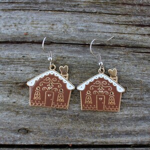 May include: A pair of gingerbread house earrings. The earrings are brown with white trim and gold accents. Each house has a heart-shaped door and a chimney with a heart-shaped top. The earrings hang from silver-colored hooks.