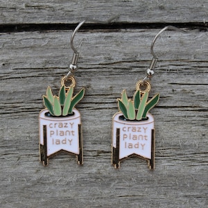 May include: Dangle earrings with a white planter design, green plant details, and the text "crazy plant lady." The earrings have gold-coloured accents and silver-coloured ear wires. The earrings are set against a weathered wood background.
