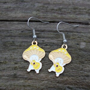 May include: A pair of whimsical mushroom-shaped earrings. Each earring features a yellow and tan mushroom cap with a smiling face and a small guitar. The earrings have silver-colored hooks and are set against a wooden background.