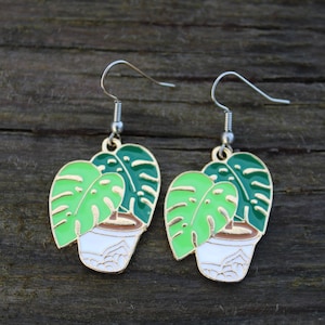 May include: A pair of earrings with a monstera plant design. Each earring features green and light green leaves, a white pot, and brown soil. The earrings have silver-coloured hooks.