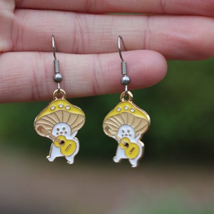 May include: A pair of whimsical mushroom-shaped earrings. Each earring features a yellow and white mushroom design with a small figure playing a yellow guitar. The earrings have silver-colored hooks and gold-colored accents.